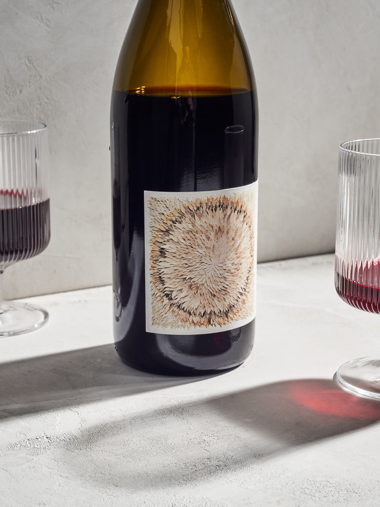 A bottle of red wine with a textured, abstract label is flanked by two half-filled glasses of wine on a light background. The scene is calm and minimalist.
