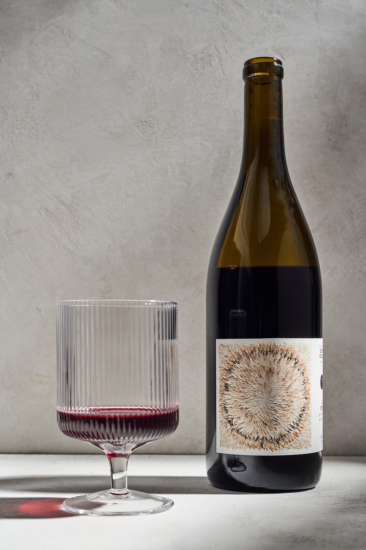 A bottle of red wine with an artistic label next to a half-full, textured glass on a neutral background. The scene exudes elegance and simplicity.