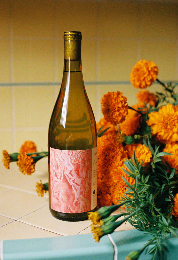 A wine bottle with a pink marbled label stands on a tiled surface surrounded by vibrant orange marigolds, set against a yellow-tiled backdrop.