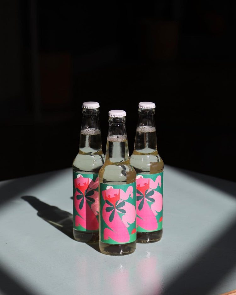 Three glass bottles with colorful labels, featuring pink and green abstract designs, stand on a light gray surface under bright, focused lighting.