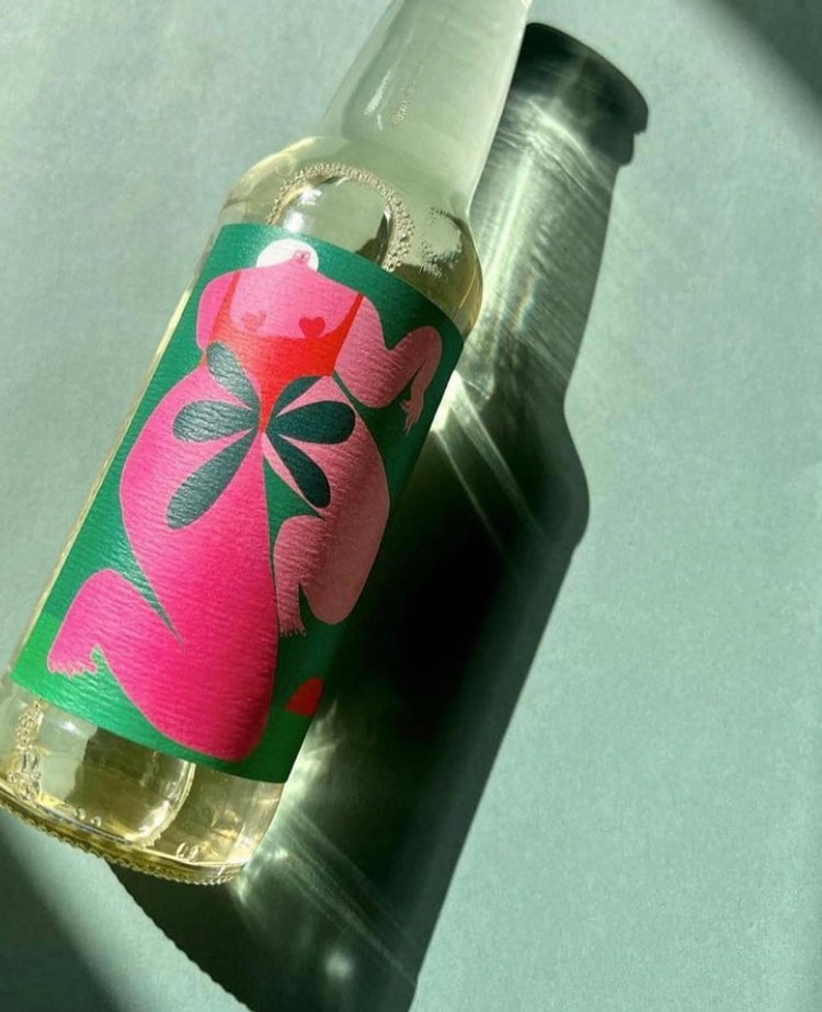 A clear glass bottle with a colorful label depicting abstract pink and green shapes casts a shadow on a green surface. The scene is bright and artistic.