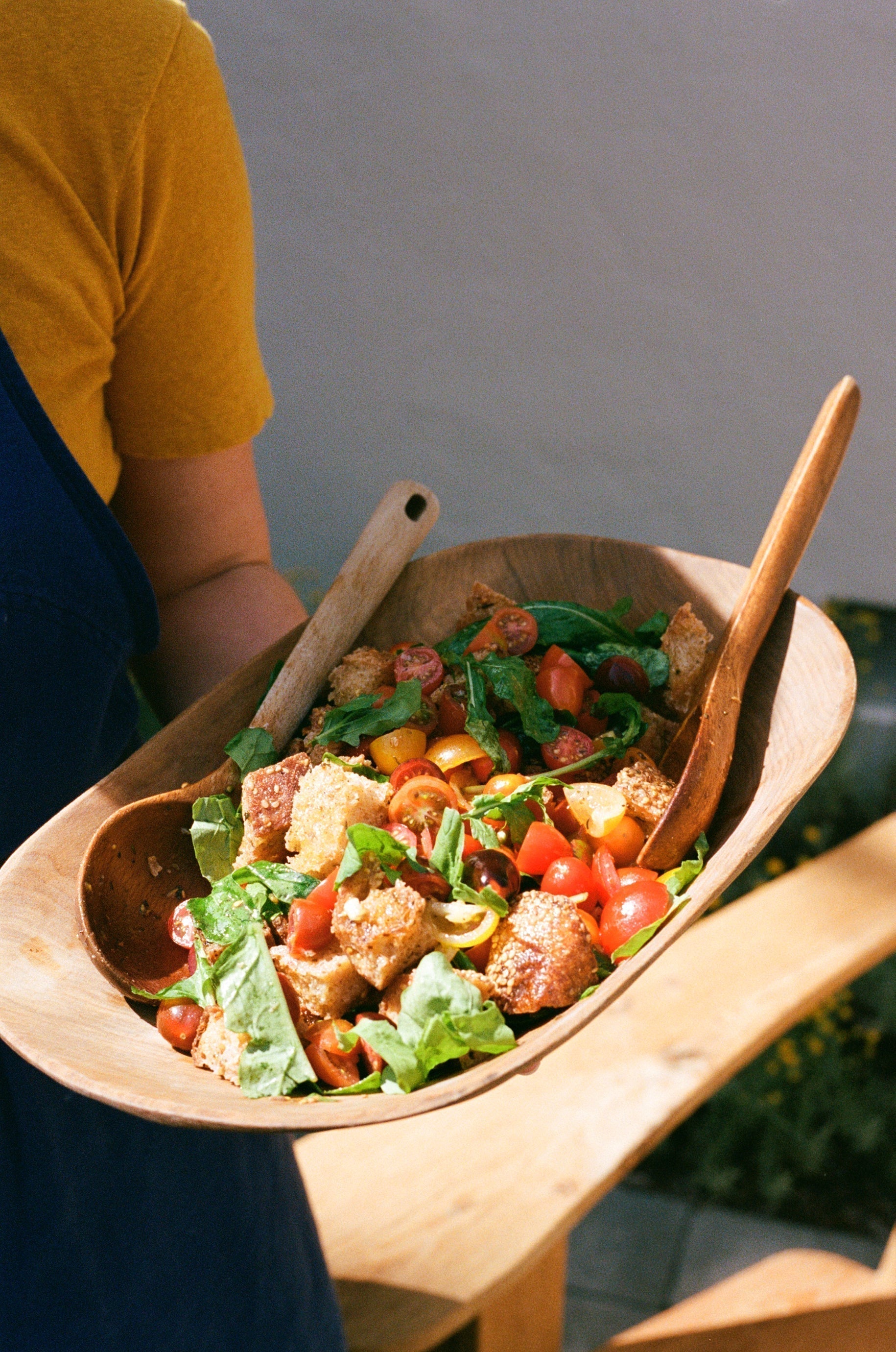 A woman in a yellow tee shirt holds a wooden bowl full of panzanella—chopped tomatoes, bread, arugula, and tongs.