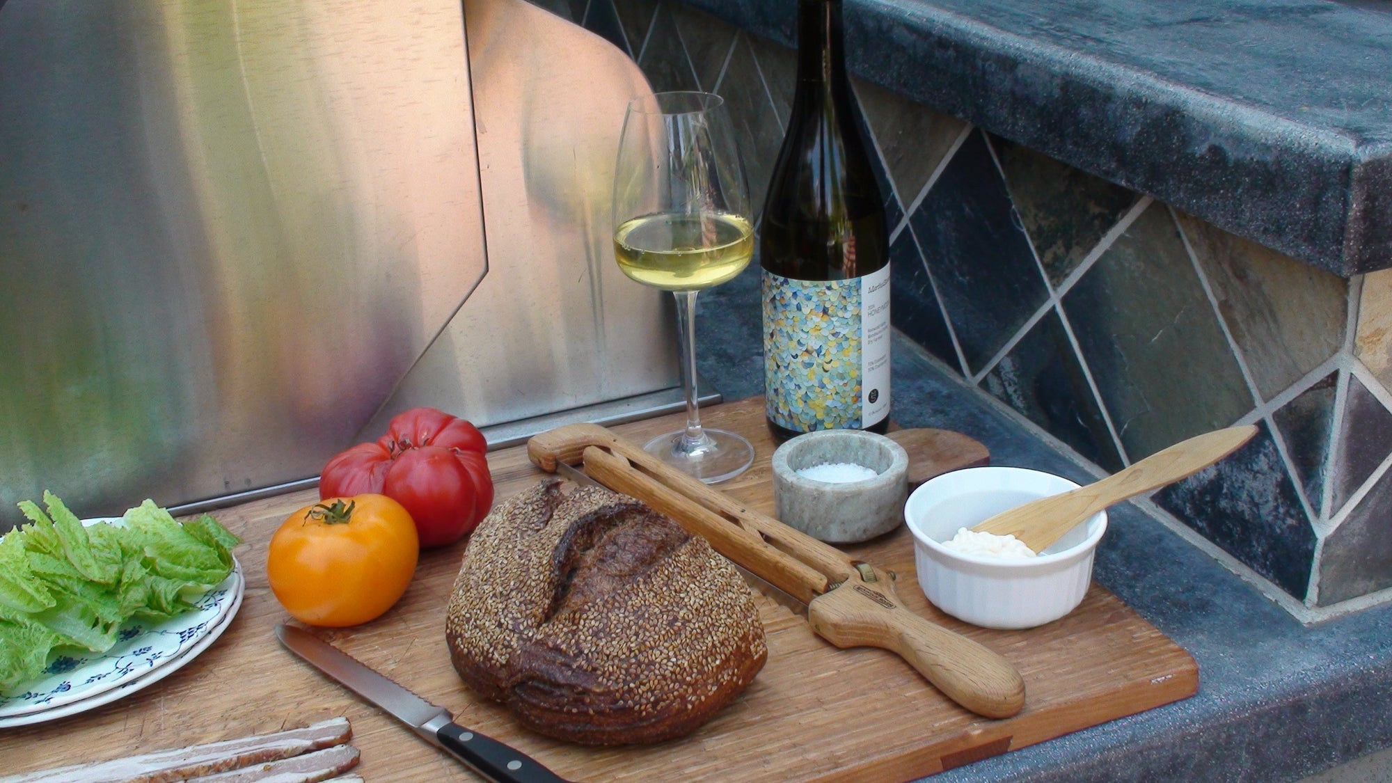 A cutting board with bread, tomatoes, salt, lettuce, and a bottle of wine sit next to a bbq on an outdoor countertop.