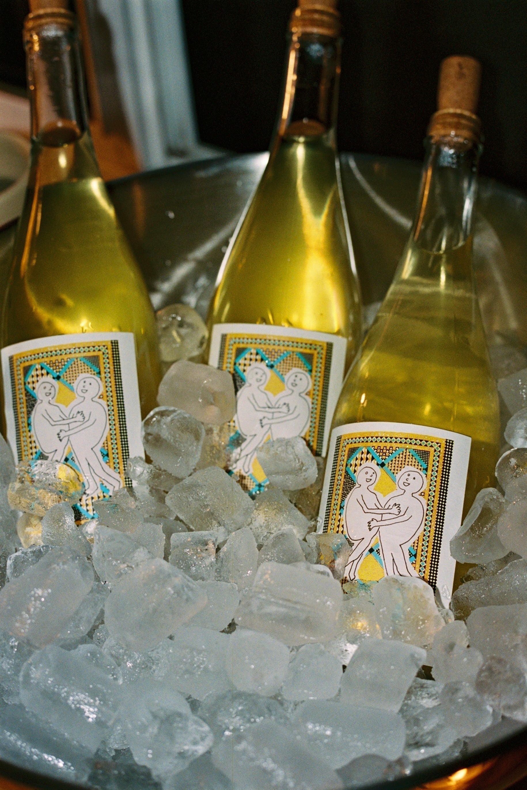 Three bottles of Post Flirtation White sit in an ice bucket. The bottles are clear glass with a yellow-hued liquid inside. The label features two  cartoonish figures hugging on a blue and yellow rug. 