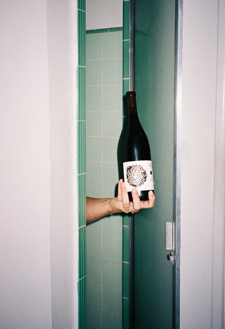 A hand extends from a shower, holding a wine bottle with a textured beige label. The bathroom features green tiles, creating a quirky, humorous tone.