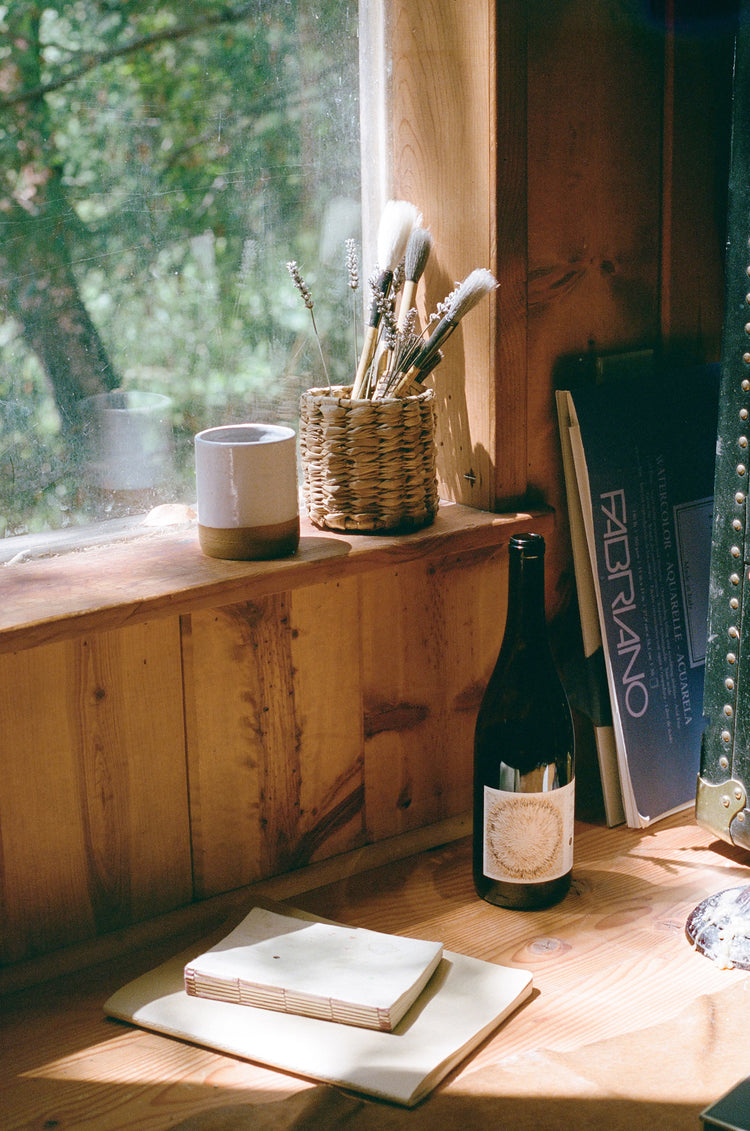 A cozy wooden windowsill with a woven basket of brushes, a ceramic cup, a wine bottle, and a stack of notebooks.