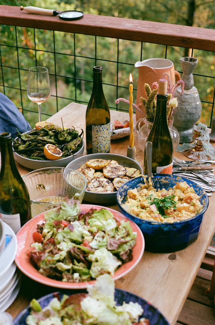 A beautifully arranged outdoor dining table with various dishes, wine bottles, and a candle, surrounded by greenery.