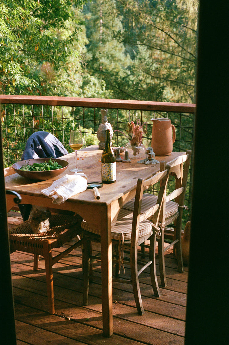 A wooden table set on a balcony, featuring a bowl of greens, a bottle of wine, and a glass, surrounded by lush greenery.