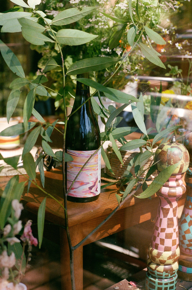 A wine bottle with an abstract label stands on a wooden table surrounded by green leaves and colorful decor items.