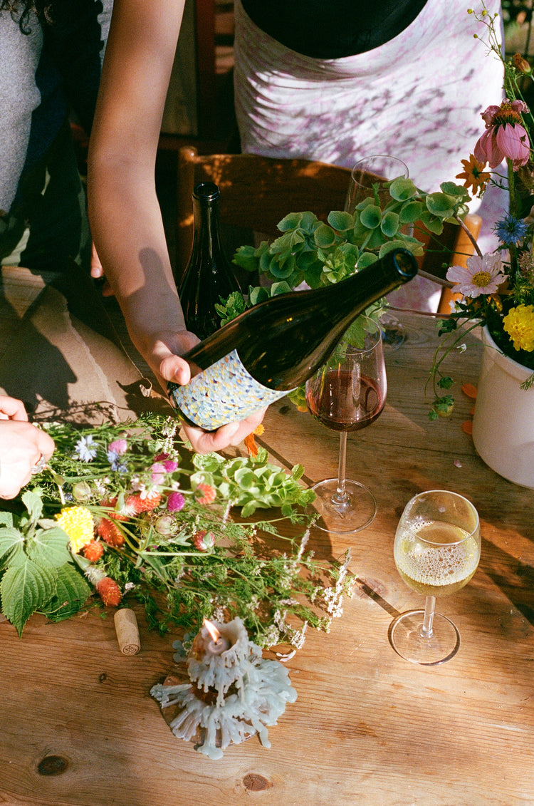 A hand pours wine from a bottle onto a table adorned with flowers, glasses, and a lit candle.