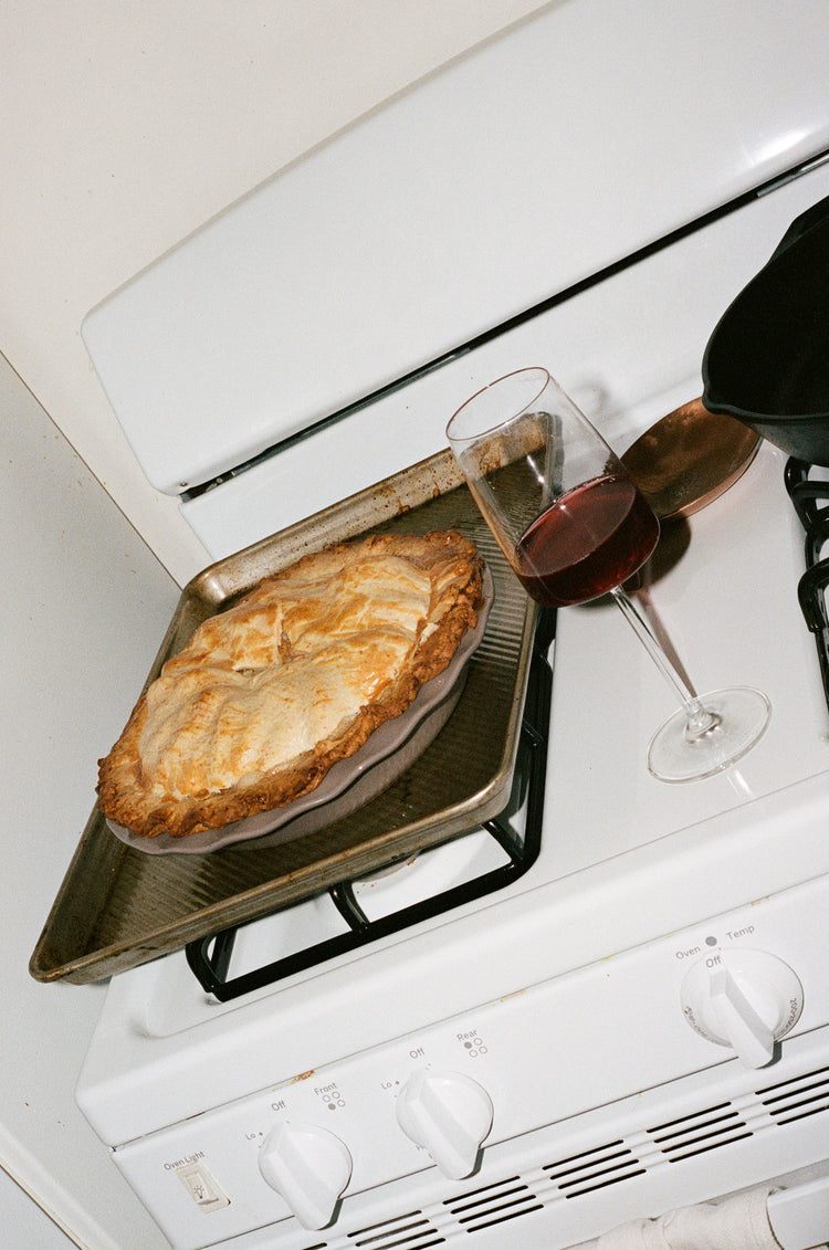 A glass of red wine on an over with a pie that has been freshly baked sitting on top of the burners cooling.