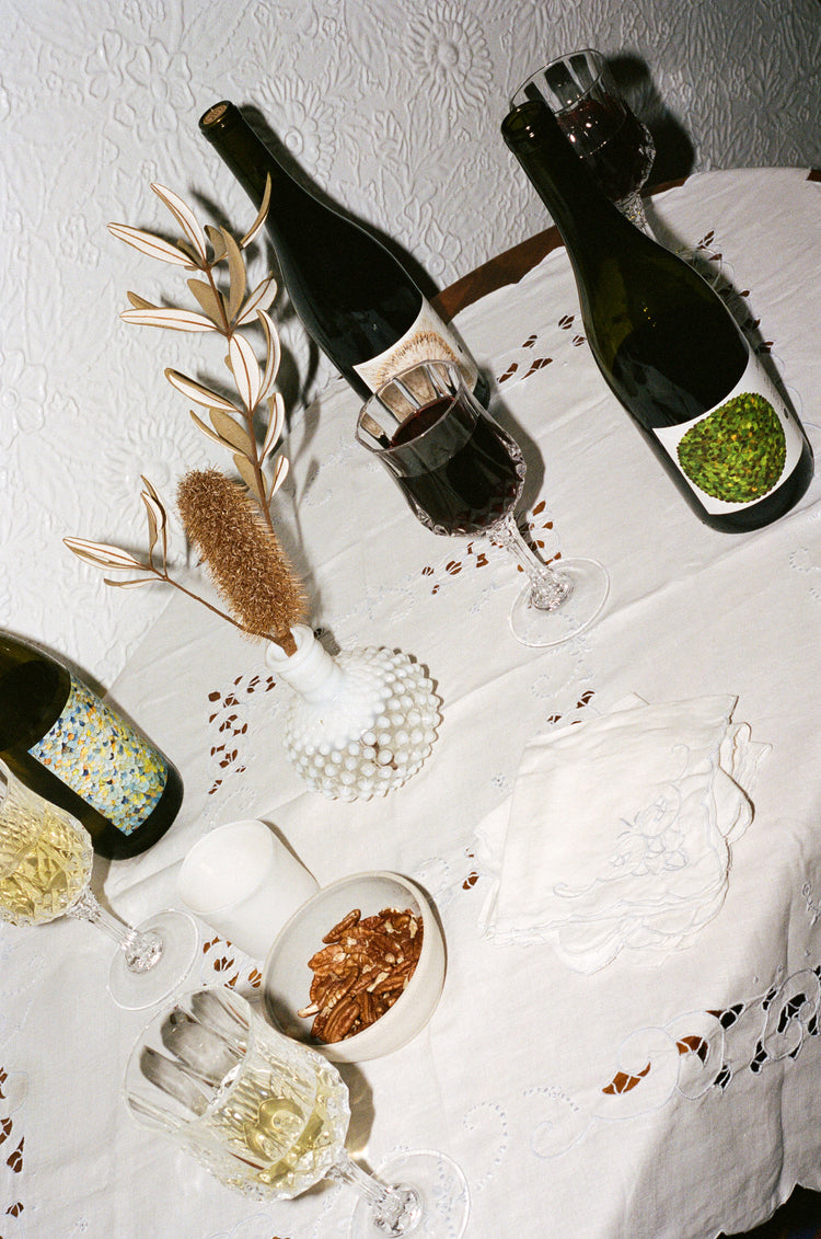 A table with a white tablecloth with three bottles of wine and four glasses of wine.