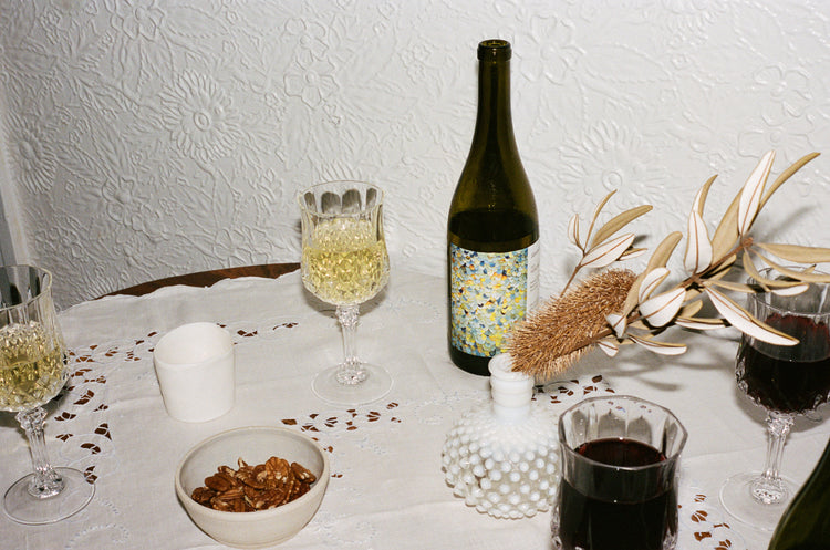 A table with a white tablecloth with four glasses of wine, a bottle of wine open sitting on the table and some walnuts in a bowl.