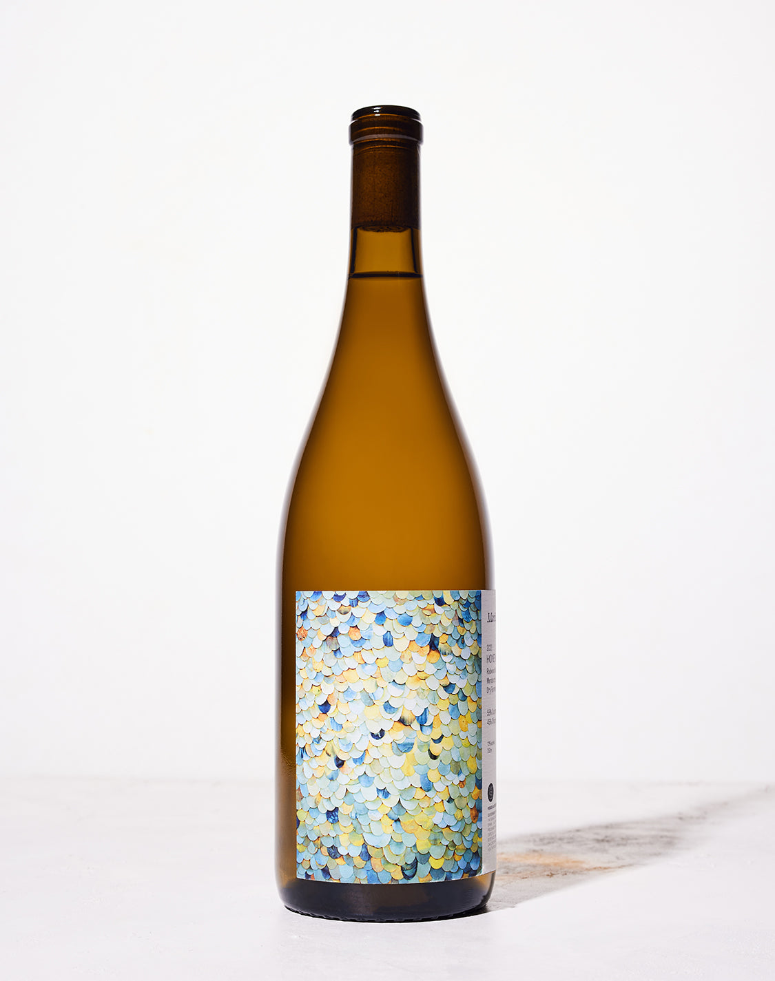 A wine bottle against a white background features a colorful abstract label with overlapping scales in shades of blue, yellow, and orange, conveying an artistic and vibrant tone.