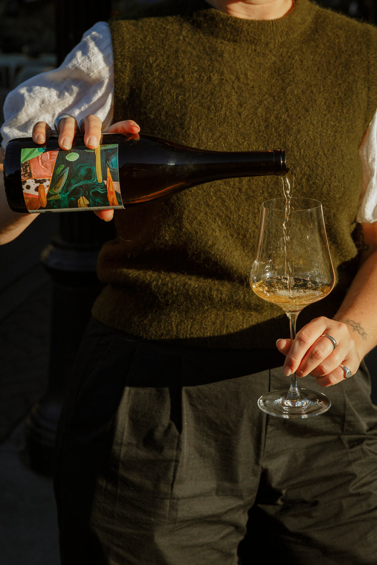 a woman in a green sweater vest holds a bottle of wine, pouring into a glass.