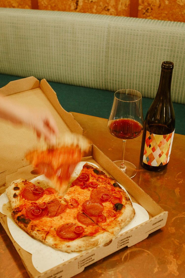 A bottle and glass of wine sit on a copper table top next to a pizza box, with a blurred hand removing a slice of pizza. 