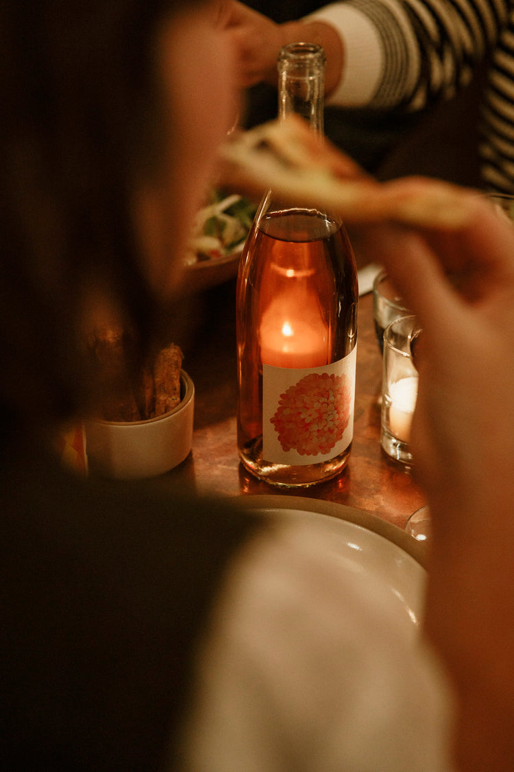 A beautiful bottle of rosé wine is illuminated from behind via candlelight. The frame is abstracted in the foreground by someone taking a bite of food.