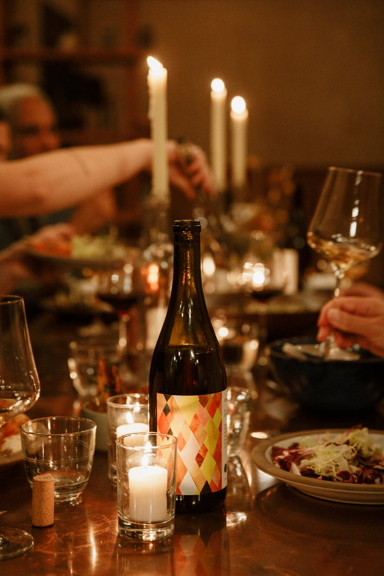 A wine bottle with a warm toned geometric label sits on a dinner table at a party, with a votive candle in front and tall candlesticks behind. 