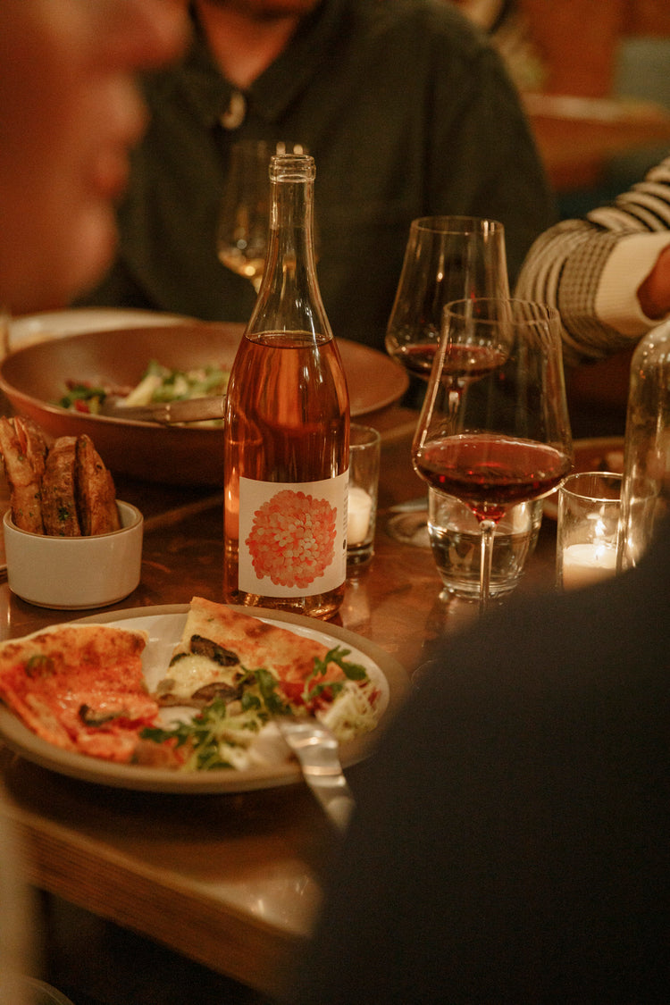 A bottle of rosé wine with a pink circular label sits on a dinner table with plates and bowls of food. 
