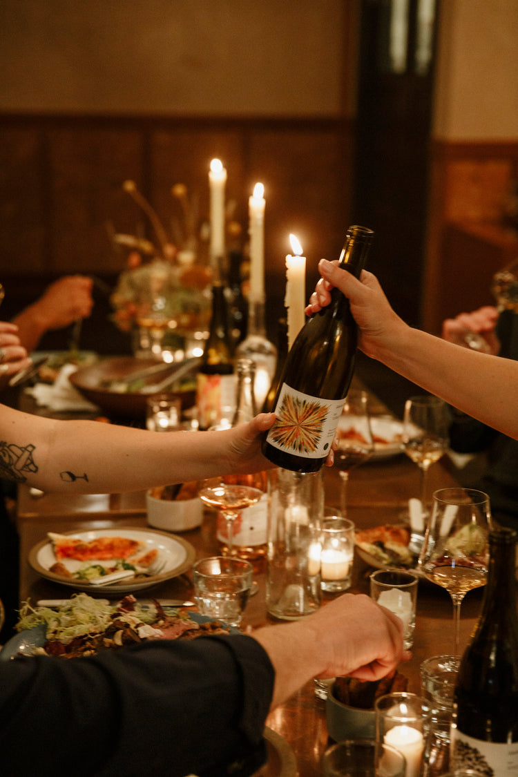 A bottle of Martha Stoumen Vermentino is passed across a dinner table in a warm, candle lit room