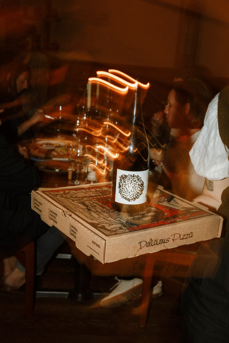 A photo of a bottle of wine with a black round logo sits on top of a pizza box, being carried by someone with tattoos. the slow shutter speed photo creates orange streaks on the frame. 