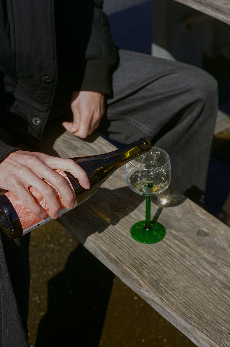 A man holds a bottle of Solo Act white wine and pours it into a small, green stemmed wine glass. The glass is on a wooden bench and the man is wearing grey jeans and a black quilted jacket. They're outside.