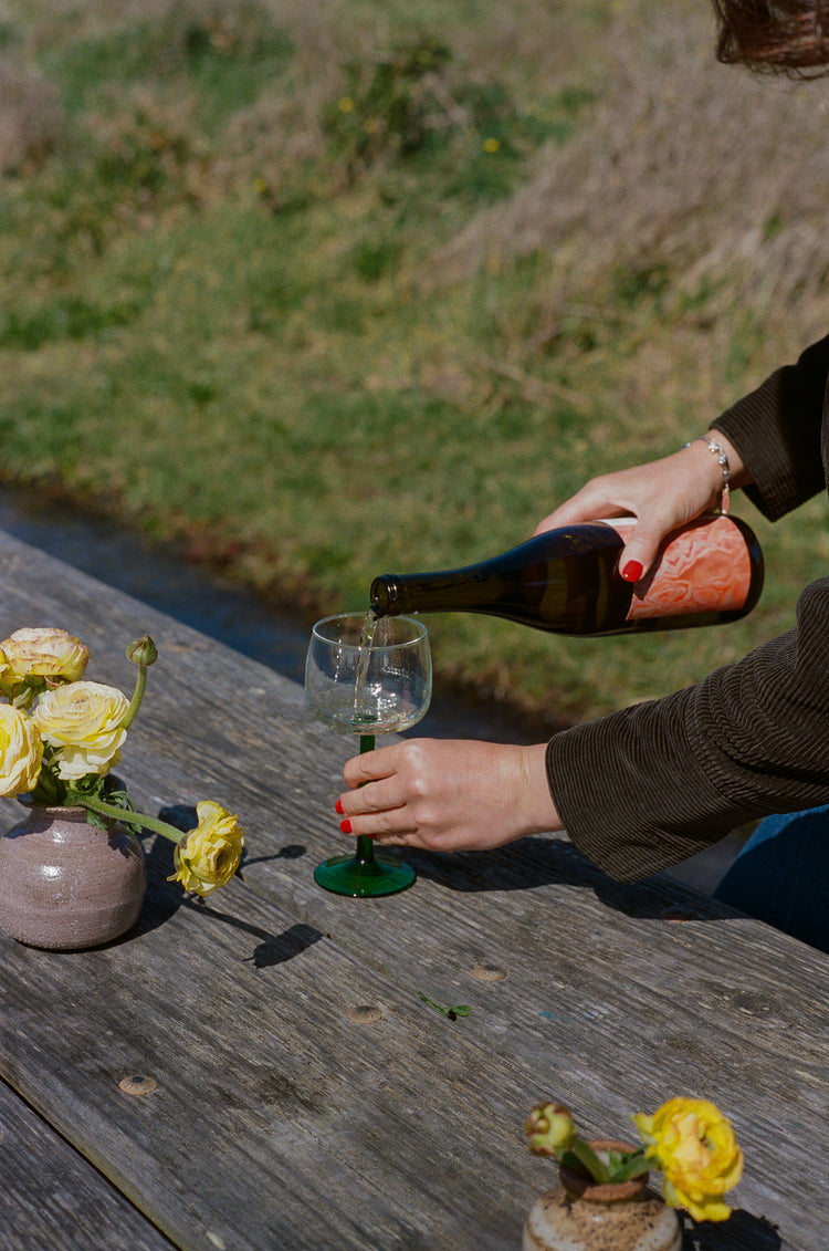 A person pours wine into a green-stemmed glass on a wooden table outdoors. Yellow flowers in a vase add a cheerful, rustic touch.