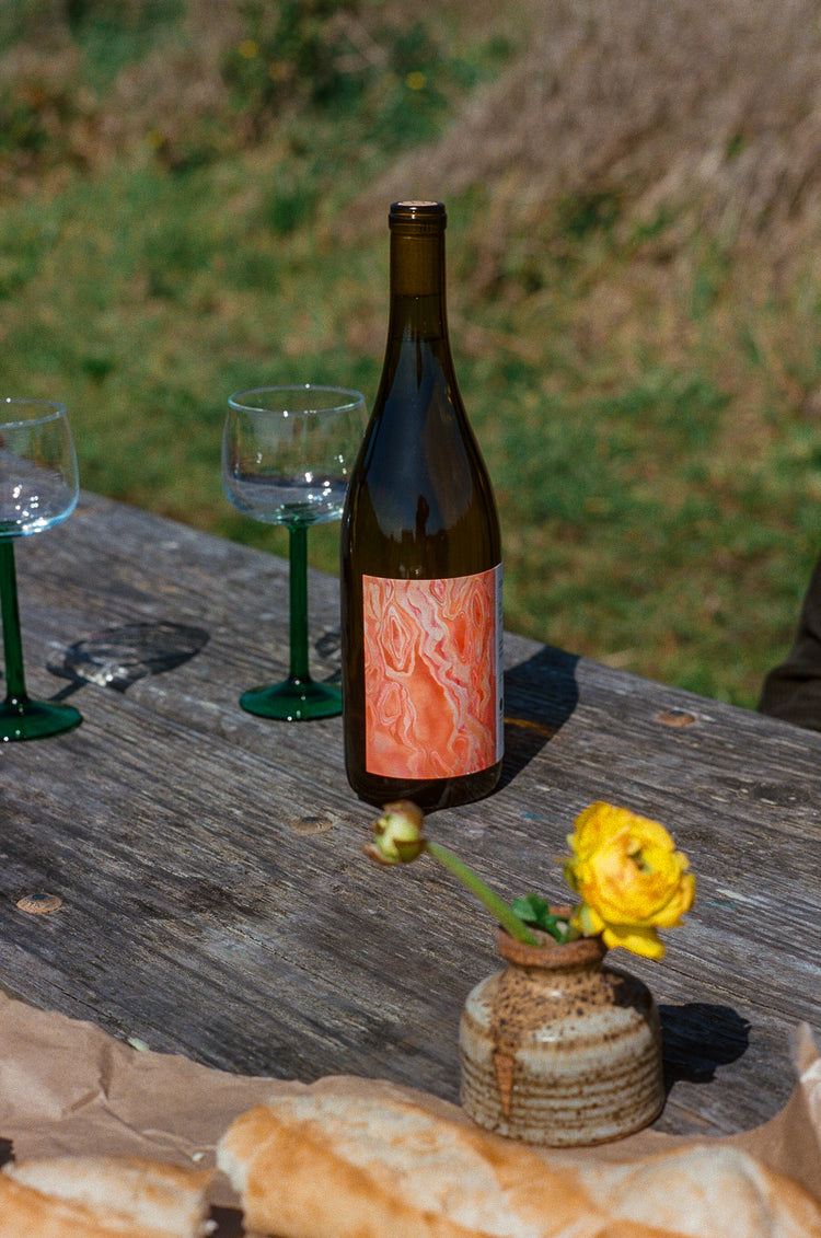 A bottle of Martha Stoumen Solo Act wine, which is a dark glass bottle with a pink and orange label, sits on a picnic table with two wine glasses and a small bud vase filled with yellow flowers. In the foreground there is a loaf of bread and in the background is greenery. 