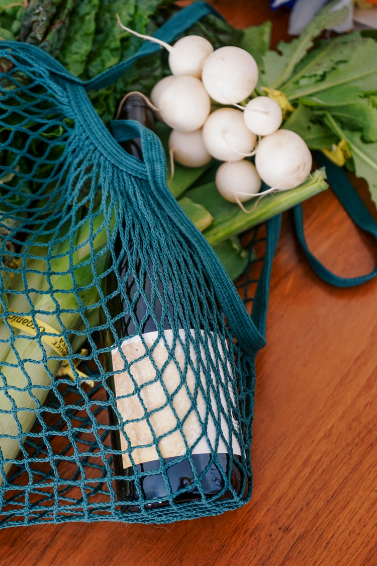 Net bag with white radishes, green vegetables, and a wine bottle on a wooden table. The scene suggests a fresh, organic market vibe.