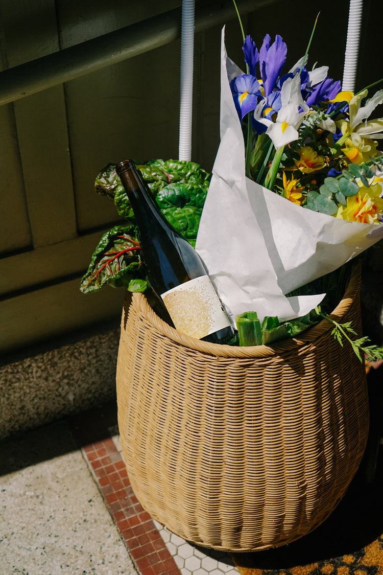 A wicker basket holds a wine bottle, colorful flowers, and leafy greens, set against a wooden wall. Sunlight casts a warm, inviting glow.