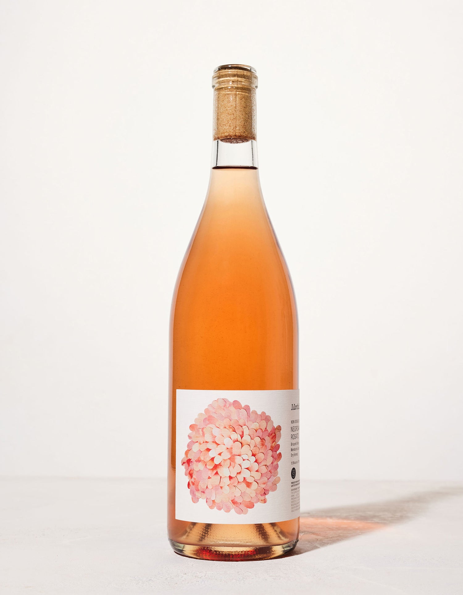 A tall bottle of rosé wine with a cork, featuring a label adorned with a cluster of pink petals. The background is a clean, soft white.