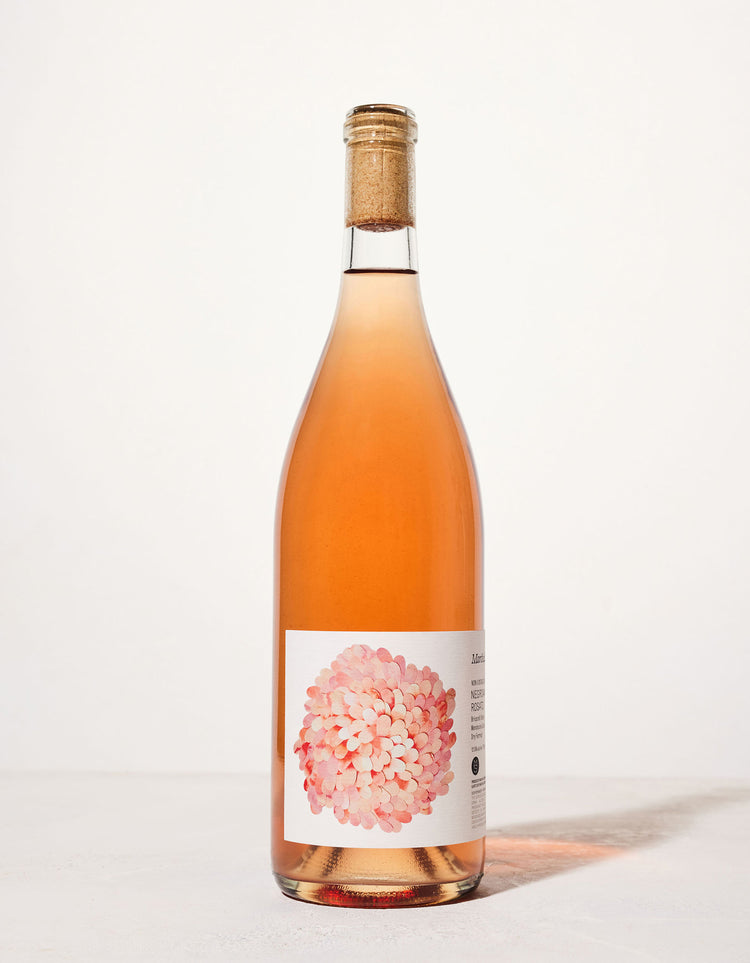 A tall bottle of rosé wine with a cork, featuring a label adorned with a cluster of pink petals. The background is a clean, soft white.