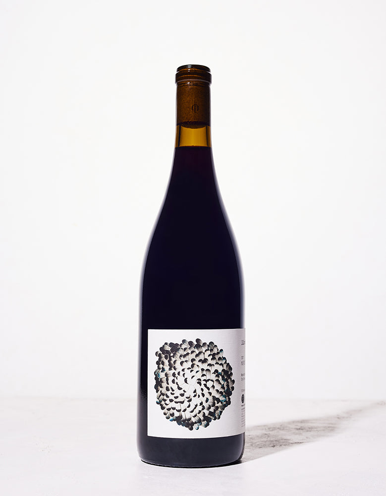 A dark wine bottle with a sleek design stands on a light surface, featuring an artistic label depicting a cluster of abstract black and blue shapes.