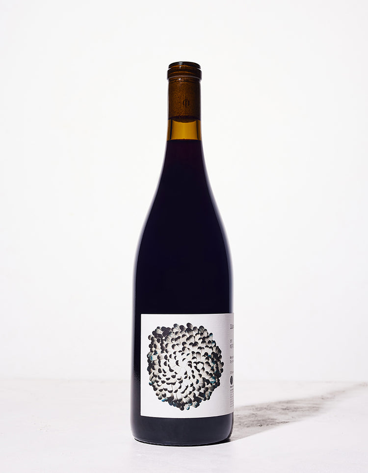 A dark wine bottle with a sleek design stands on a light surface, featuring an artistic label depicting a cluster of abstract black and blue shapes.
