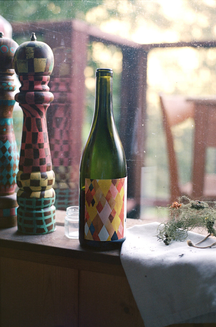 A green wine bottle with a colorful diamond-patterned label stands on a sunlit windowsill beside tall, patterned salt and pepper grinders, evoking a cozy, rustic ambiance.