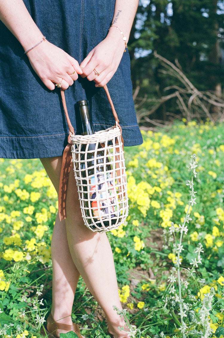A woman in a denim dress holds a woven basket with a bottle of wine inside.