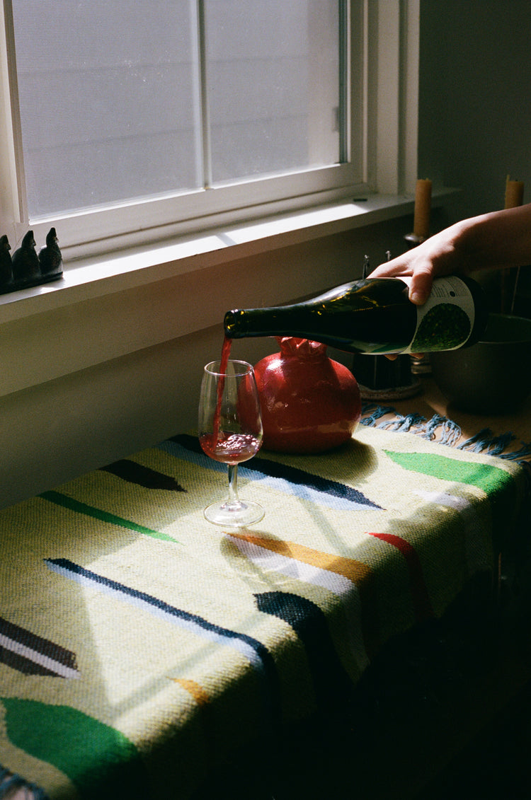 Person pouring wine into a glass on a colorful woven tablecloth with a window in the background