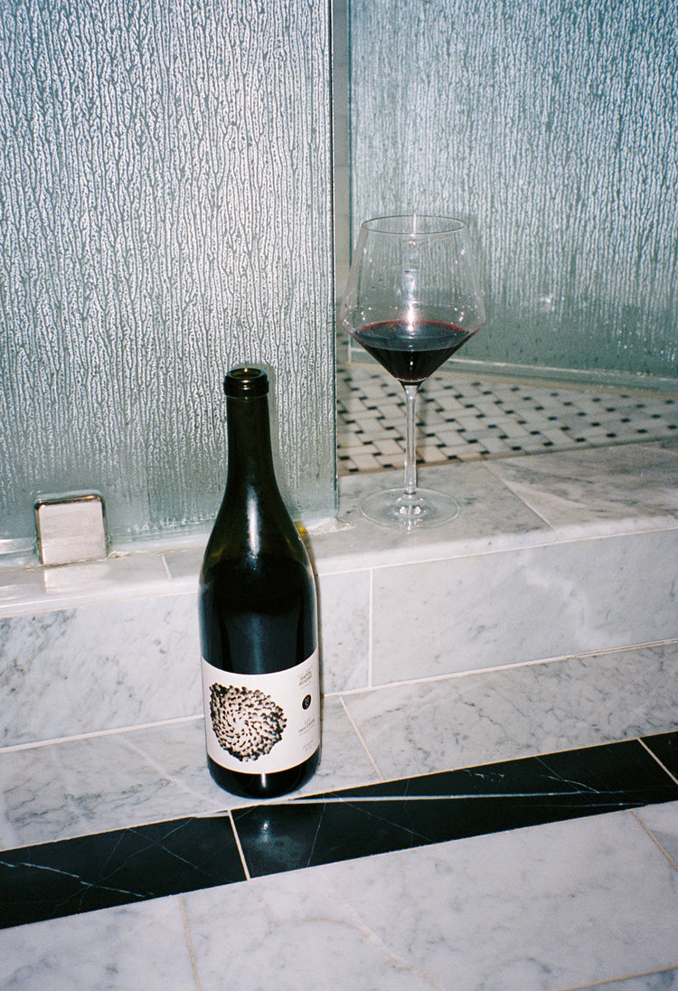 A wine bottle and glass of red wine are placed on a marble ledge beside a textured glass wall. The scene conveys a sense of luxury and relaxation.