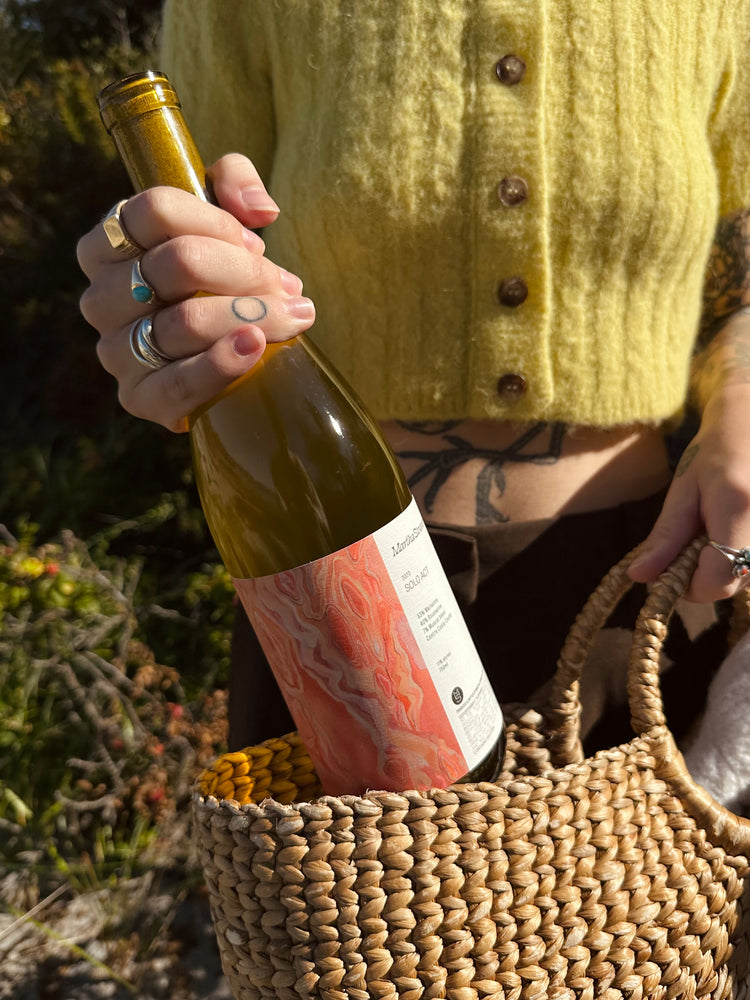 A person in a yellow sweater, wearing rings, placing a wine bottle with a marbled label into a woven straw bag. Sunlit scene with a relaxed vibe.