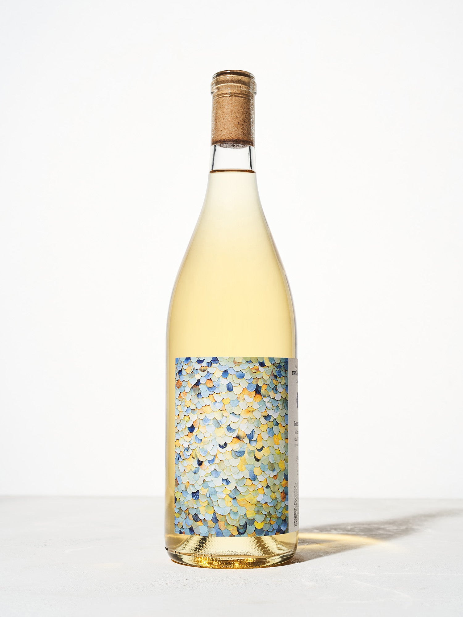 A wine bottle with a cork features a colorful label with a pattern of overlapping scales in blue, yellow, and orange hues. Neutral background.