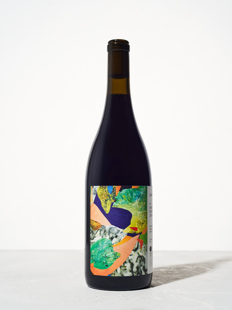 A wine bottle with a colorful abstract label featuring vibrant greens, blues, and oranges. The background is a smooth, light gray surface.