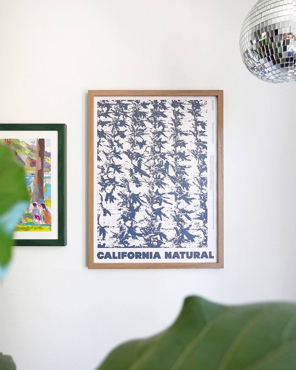 Wall art scene with a poster titled "California Natural," featuring abstract blue foliage. A leafy plant and a disco ball add a lively touch.