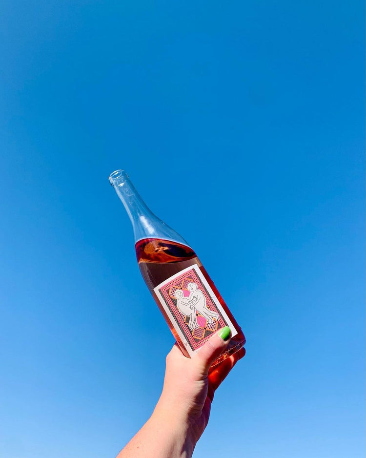A hand with green nail polish holds a clear bottle of rosé wine against a bright blue sky. The label features two abstract figures dancing.