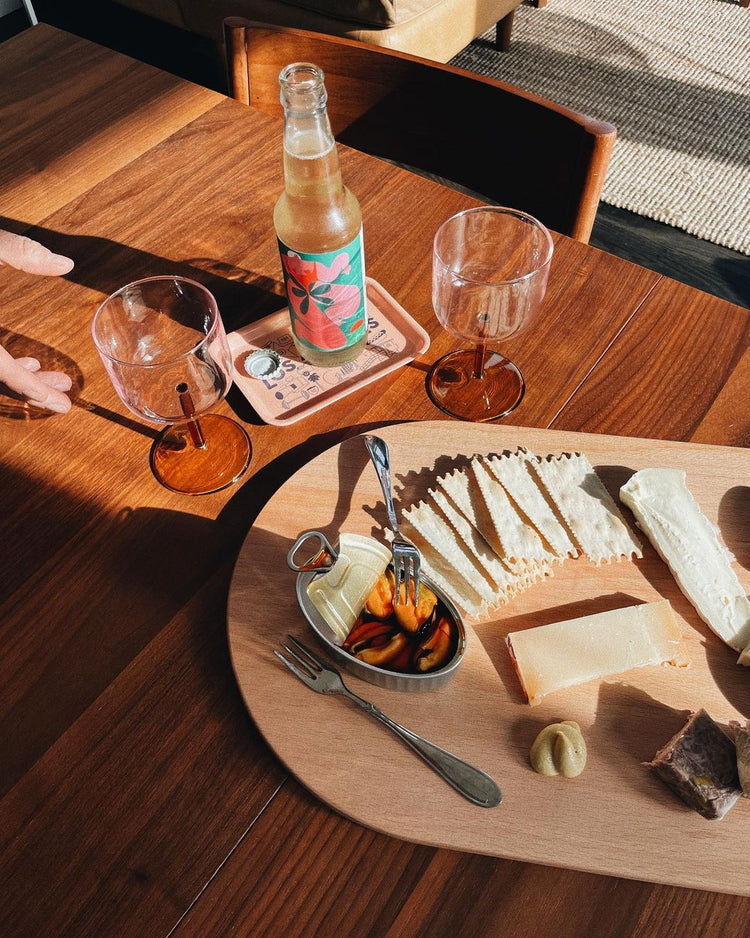 A wooden table with a cheese board featuring crackers, cheese, mustard, and canned seafood. A bottle and two empty glasses are nearby, bathed in warm sunlight.